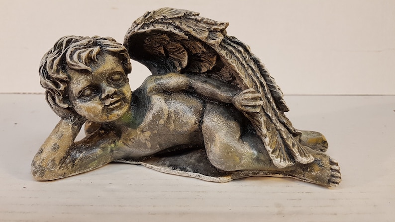 Peut inclure: Une petite figurine de ch&eacute;rubin ail&eacute; grise avec une finition vieillie. Le ch&eacute;rubin est couch&eacute; sur le c&ocirc;t&eacute;, la t&ecirc;te reposant sur sa main. Les ailes sont d&eacute;ploy&eacute;es derri&egrave;re le ch&eacute;rubin.