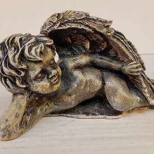 Peut inclure: Une statue de ch&eacute;rubin en pierre grise avec des ailes, couch&eacute;e sur le c&ocirc;t&eacute;. Le ch&eacute;rubin a une expression douce et d&eacute;licate sur son visage.