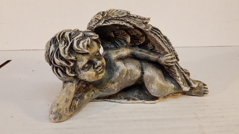 Peut inclure: Une statue de ch&eacute;rubin en pierre grise avec des ailes, couch&eacute;e sur le c&ocirc;t&eacute;. Le ch&eacute;rubin a une expression douce et d&eacute;licate sur son visage.