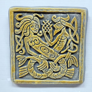 May include: A ceramic wall plaque featuring a detailed relief sculpture of two intertwined mermaids. The mermaids are depicted in a stylised, Celtic knotwork design with gold accents against a grey background.