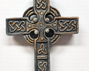 Może przedstawiać: Dekoracja ścienna w kształcie celtyckiego krzyża z misternymi wzorami węzłów w kolorze złotym i czarnym. Krzyż wykonany jest z fakturowanego materiału i ma trójwymiarowy wygląd.