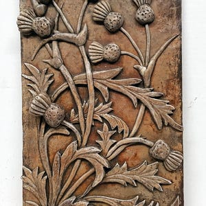 May include: A brown and silver wall sculpture featuring a raised design of stylised thistle flowers. The sculpture has a rustic, textured look.