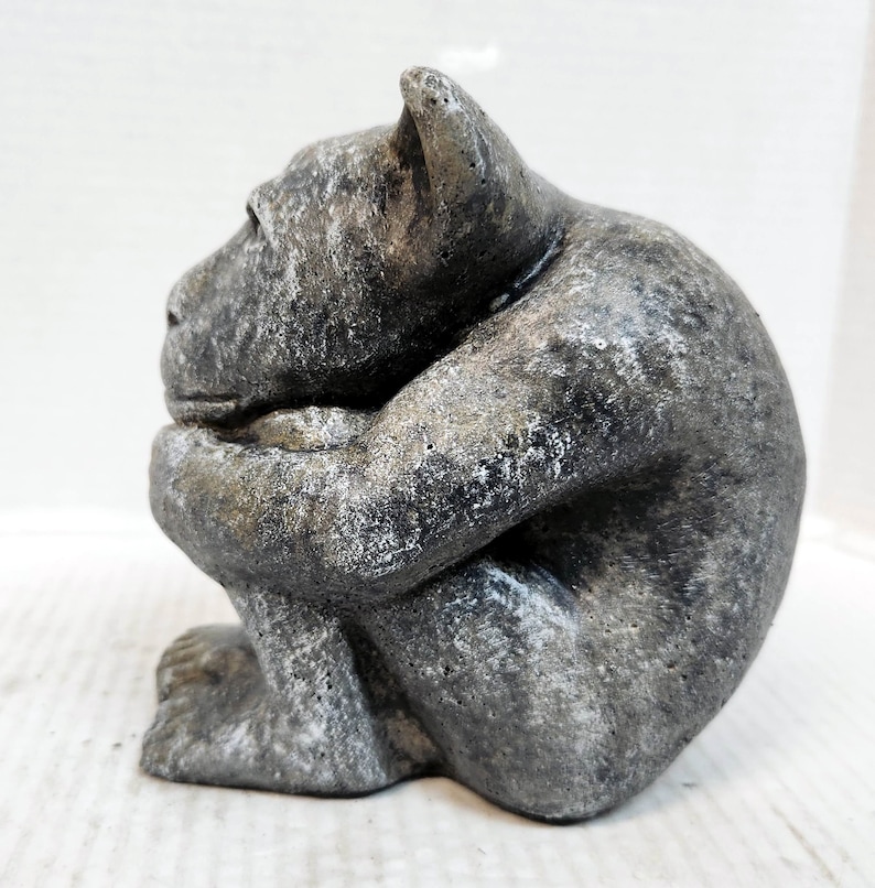 May include: A gray stone sculpture of a gargoyle sitting with its arms crossed and head down. The gargoyle has a textured surface and a weathered look.