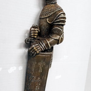 Puede incluir: Una figura de caballero de color marr&oacute;n y plateado que sostiene una espada. El caballero lleva armadura y casco. La figura est&aacute; de pie sobre una base con un dise&ntilde;o decorativo.