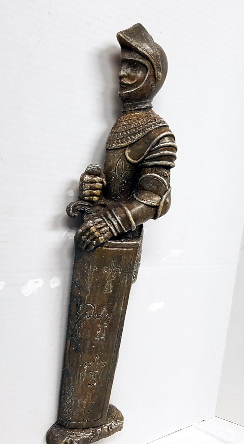 Puede incluir: Una figura de caballero de color marr&oacute;n y plateado que sostiene una espada. El caballero lleva armadura y casco. La figura est&aacute; de pie sobre una base con un dise&ntilde;o decorativo.