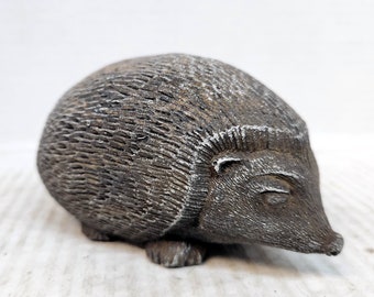 May include: A gray ceramic hedgehog figurine with a detailed textured body and a closed eye. The figurine is lying on its side with its head turned to the right.