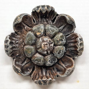 May include: A weathered, decorative flower made of a dark, possibly metallic material. The flower has multiple layers of petals with a central, textured core. The color palette includes shades of brown, green, and hints of white, suggesting an aged appearance.