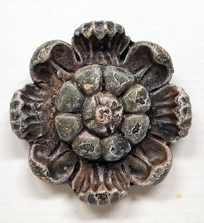 May include: A weathered, decorative flower made of a dark, possibly metallic material. The flower has multiple layers of petals with a central, textured core. The color palette includes shades of brown, green, and hints of white, suggesting an aged appearance.