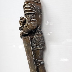 Puede incluir: Una figurita de caballero de color bronce de pie con una espada y un escudo. El caballero lleva armadura y casco. La figurita est&aacute; sobre una base de piedra.