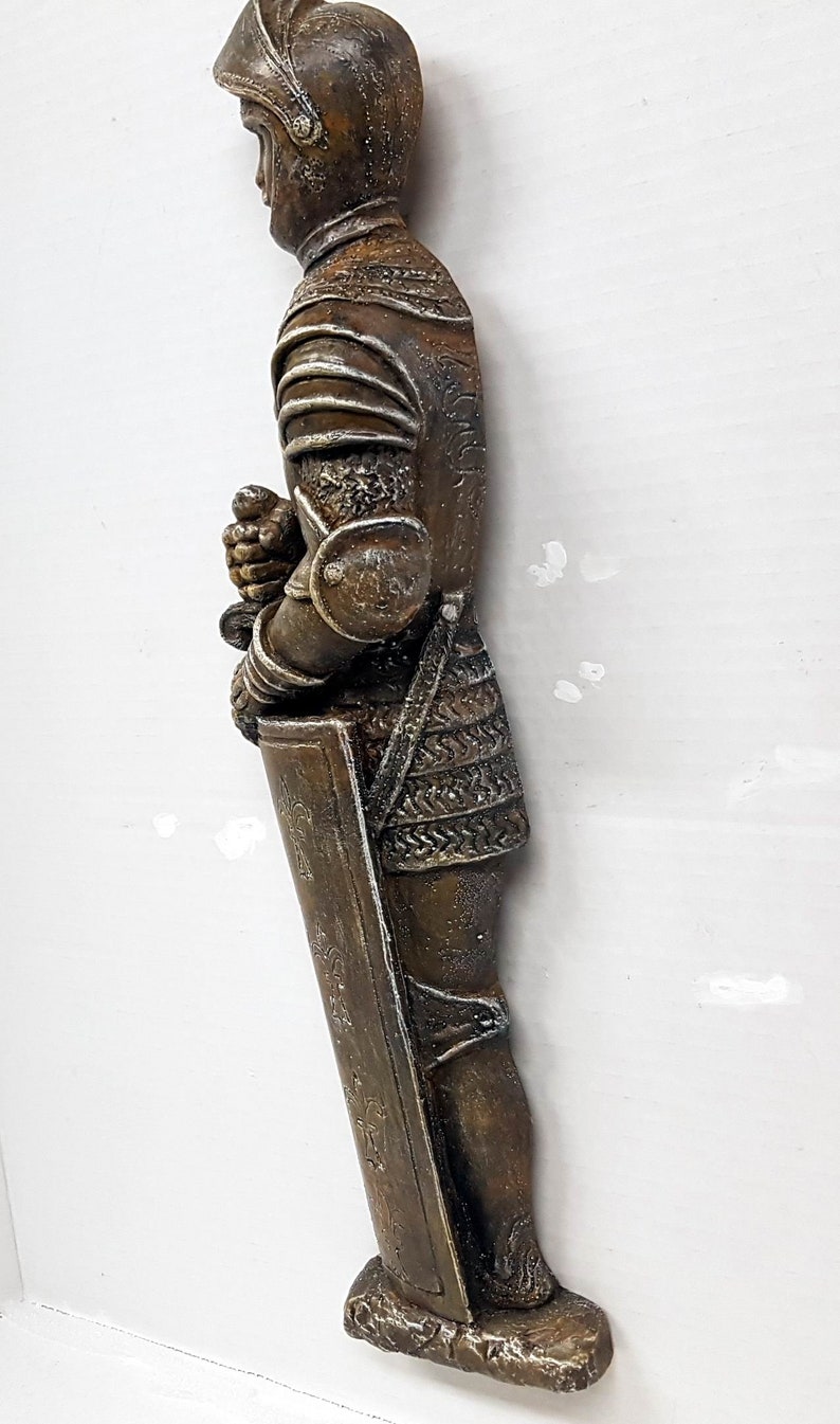 Puede incluir: Una figurita de caballero de color bronce de pie con una espada y un escudo. El caballero lleva armadura y casco. La figurita est&aacute; sobre una base de piedra.