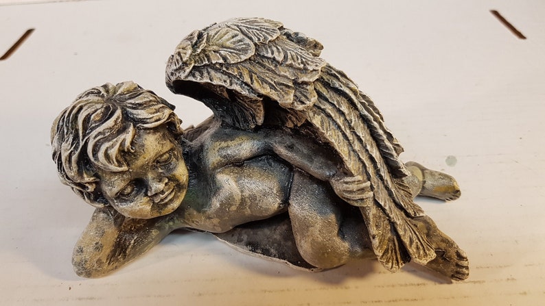 Peut inclure: Une petite figurine d'ange en pierre grise avec des ailes. L'ange est couch&eacute; sur le c&ocirc;t&eacute;, la t&ecirc;te reposant sur son bras. L'ange a une expression douce et innocente sur son visage.