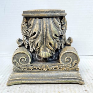 May include: A decorative, ornate, gold-toned, plaster column base with a detailed, leaf-like design. The base has a square platform and a rounded top.