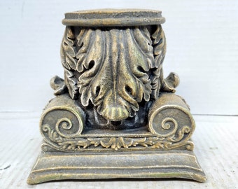 Può includere: Una base di colonna decorativa in gesso, color oro, decorata con un designo dettagliato a forma di foglie. La base ha una piattaforma quadrata e una parte superiore arrotondata.