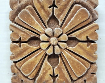 May include: A square, decorative tile with a raised, intricate design. The tile is made of a light brown material and features a central flower with eight petals. The flower is surrounded by a repeating pattern of curved lines and shapes.