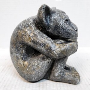 May include: A gray stone gargoyle sculpture with a distressed finish. The gargoyle is sitting with its arms crossed and its head tilted down.