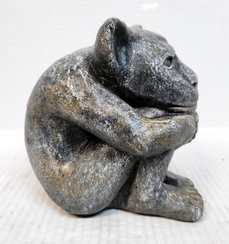 May include: A gray stone gargoyle sculpture with a distressed finish. The gargoyle is sitting with its arms crossed and its head tilted down.
