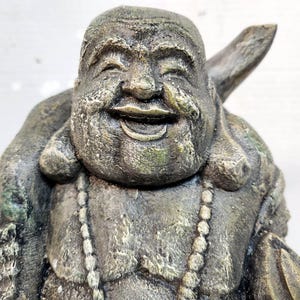 May include: A weathered, stone-like statue of a smiling Buddha. The statue has a detailed face, a beaded necklace, and a large ear. The color is a mix of gray, green, and brown, giving it an aged appearance. The Buddha is depicted with a serene expression.