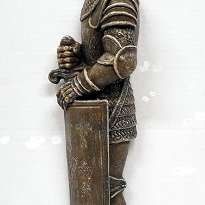 Puede incluir: Una figurita de bronce de un caballero con armadura sosteniendo una espada y un escudo. El caballero lleva un casco con visera y una t&uacute;nica de malla. El escudo est&aacute; decorado con flores de lis.