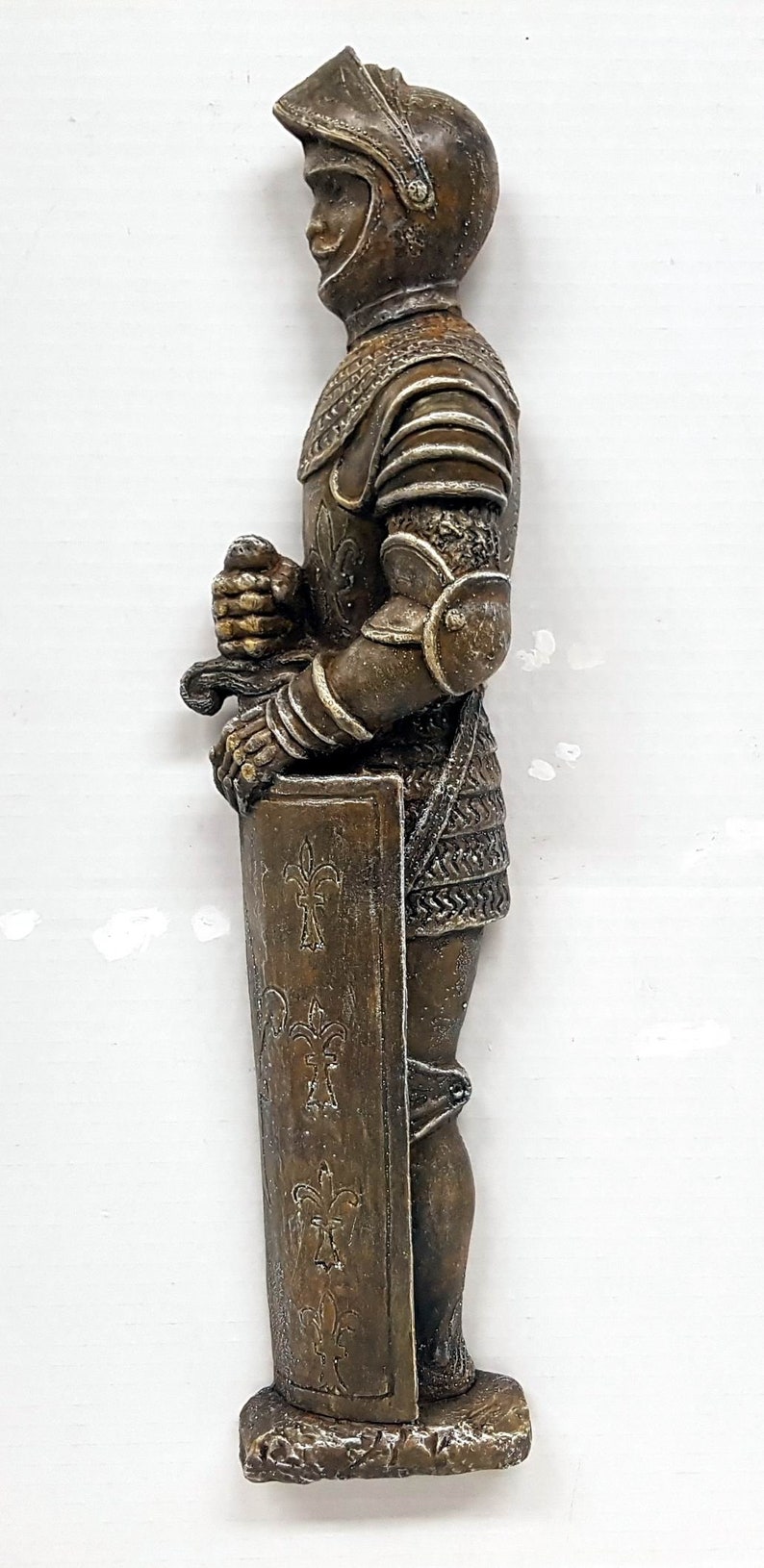 Puede incluir: Una figurita de bronce de un caballero con armadura sosteniendo una espada y un escudo. El caballero lleva un casco con visera y una t&uacute;nica de malla. El escudo est&aacute; decorado con flores de lis.