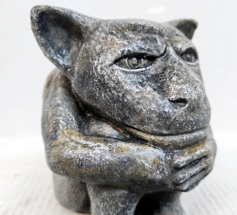 May include: A gray stone gargoyle sculpture with a detailed face and a hunched posture. The gargoyle has a furrowed brow, a prominent nose, and a closed mouth.