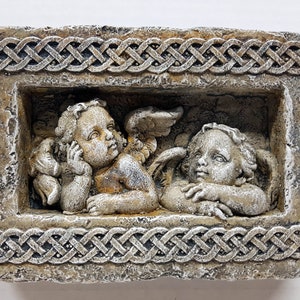 May include: A stone wall plaque with a Celtic knot border, featuring two cherub angels with wings. The angels are sculpted in a light brown and white color.