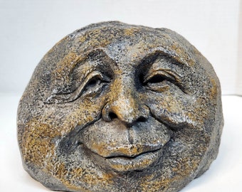 May include: A whimsical stone sculpture of a face with a wide, toothy grin. The face is sculpted in a realistic style, with detailed features and a textured surface. The stone is a mix of gray and brown, with hints of gold.
