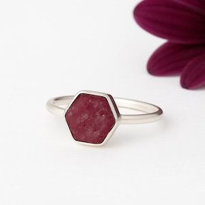以下が含まれることがあります： 六角形の濃い赤色の石が付いたシルバーリング。石はテクスチャ加工された表面を持ち、シルバーのベゼルにセットされています。リングは白い背景にあり、背景にはぼやけた紫色の花があります。