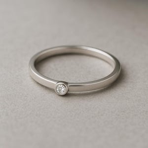 May include: A silver ring featuring a small, round, clear gemstone in a bezel setting. The ring band is plain, polished metal. The ring is set against a neutral grey background, emphasising its minimalist design. This ring is a piece of jewellery.