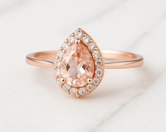 May include: Rose gold ring featuring a pear-shaped peach-colored gemstone surrounded by small, clear accent stones. The ring has a delicate band and a halo design, set against a white marble background.