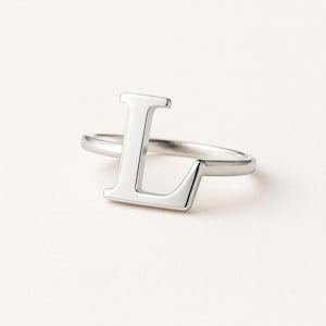 May include: A silver-colored ring featuring the capital letter "L". The ring is made of a shiny, polished metal and sits on a white surface. The letter "L" is the primary design element, standing out prominently.