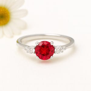 Op de afbeelding: Een zilveren ring met een grote, ronde, helderrode edelsteen in het midden, geflankeerd door twee kleinere, heldere edelstenen aan weerszijden. De ring staat tegen een witte achtergrond, met een madeliefje op de achtergrond.