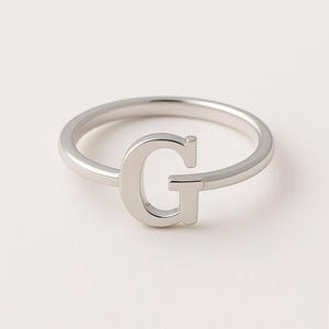 Può includere: Un anello color argento con la lettera maiuscola "G" come elemento principale. L'anello ha una fascia semplice e la lettera è visualizzata in un carattere sans-serif. L'anello è probabilmente realizzato in metallo lucido.