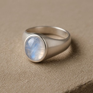 Op de afbeelding: Zilveren ring met een ovale maansteen. De maansteen heeft een blauwe en witte glans. De ring heeft een eenvoudig, modern ontwerp met een gladde, geborstelde afwerking.