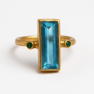 May include: A gold-toned ring featuring a large, rectangular, light blue gemstone as the centerpiece. Two small, round, green gemstones are set on either side. The ring has a simple, elegant design.