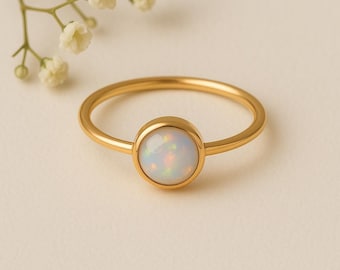 May include: A gold-colored ring with a round, iridescent opal stone. The ring band is thin and simple, and the opal has a milky white base with flashes of color. The ring is set against a soft, neutral background.