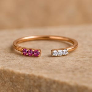 Può includere: Un anello aperto in oro rosa con tre gemme rosa da un lato e tre gemme trasparenti dall'altro. L'anello è presentato su uno sfondo neutro e strutturato.