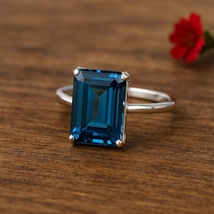 Könnte beinhalten: Ein silberner Ring mit einem großen, rechteckigen, tiefblauen Edelstein. Der Edelstein ist der Mittelpunkt, gefasst in einem schlichten, eleganten Band. Der Ring wird auf einer Holzoberfläche präsentiert, mit einer roten Blume im Hintergrund.