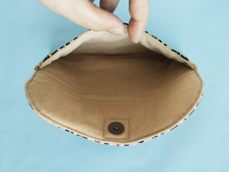 Puede incluir: Bolso de mano abierto con interior beige y exterior con estampado negro y beige. El bolso de mano se mantiene abierto, revelando el forro interior y un cierre magn&eacute;tico a presi&oacute;n. El fondo es azul claro.