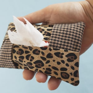 May include: A small, rectangular tissue holder with a brown leopard print and a brown and black checkered pattern. White tissues are visible. The holder is held in a hand against a light blue background.