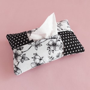 May include: A white tissue box cover with a black and white floral pattern. The cover is made of fabric and has a patchwork design. The tissue box is sitting on a pink surface.