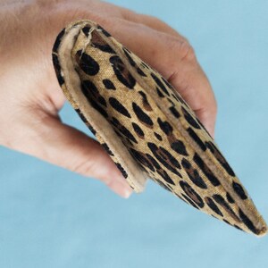 Puede incluir: Una cartera rectangular plegada con un estampado de leopardo. La cartera se sostiene en una mano sobre un fondo azul claro. El estampado de leopardo presenta manchas marrones y negras sobre un fondo beige.