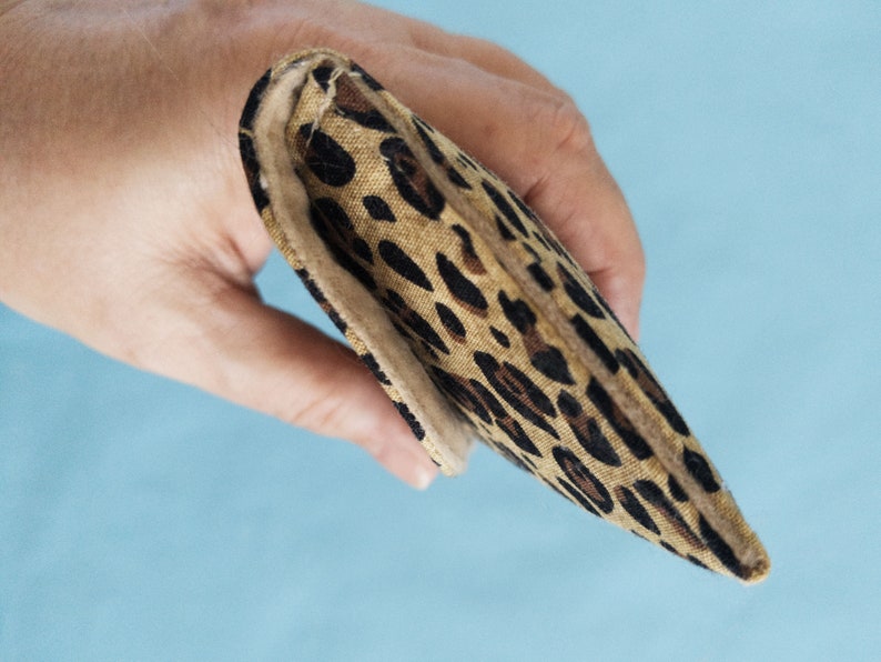 Puede incluir: Una cartera rectangular plegada con un estampado de leopardo. La cartera se sostiene en una mano sobre un fondo azul claro. El estampado de leopardo presenta manchas marrones y negras sobre un fondo beige.