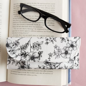 May include: A white eyeglasses case with a black floral pattern, next to a pair of black-framed glasses. The case is open, revealing a soft interior. The items rest on an open book.