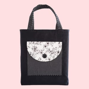 May include: A black tote bag featuring two handles with a white polka dot pattern. The front of the bag has a square pocket with a white polka dot base and a rounded flap decorated with a black and white floral print, secured by a black button.