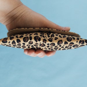 Puede incluir: Un peque&ntilde;o accesorio puntiagudo con estampado de leopardo se sostiene en una mano. El art&iacute;culo tiene un interior beige y un exterior manchado marr&oacute;n y negro. El fondo es azul claro. El accesorio parece ser una peque&ntilde;a bolsa o estuche.
