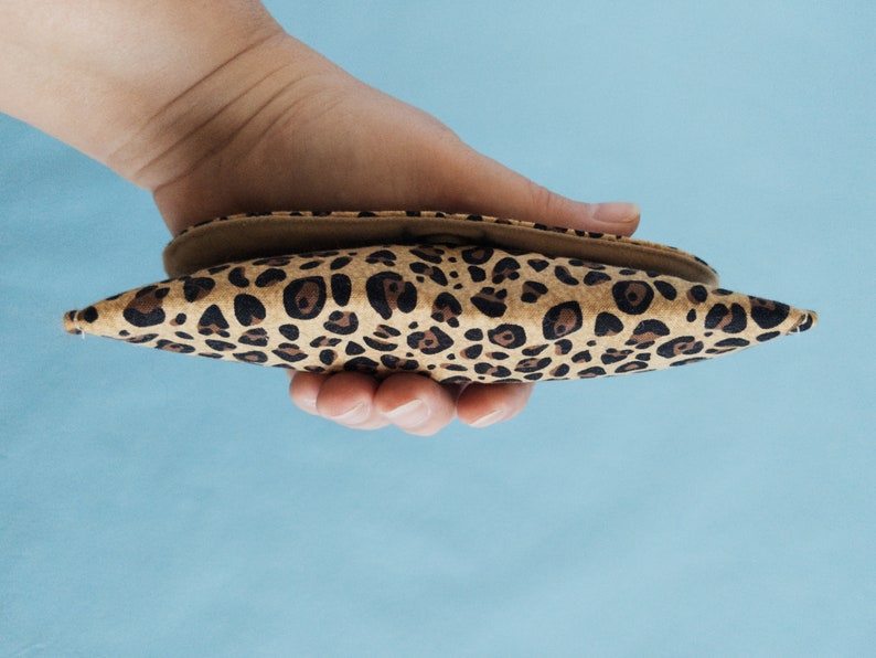 Puede incluir: Un peque&ntilde;o accesorio puntiagudo con estampado de leopardo se sostiene en una mano. El art&iacute;culo tiene un interior beige y un exterior manchado marr&oacute;n y negro. El fondo es azul claro. El accesorio parece ser una peque&ntilde;a bolsa o estuche.