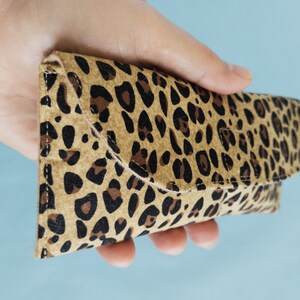 Puede incluir: Estuche para gafas con estampado de leopardo hecho de tela. El estuche se sostiene en una mano y tiene un cierre de solapa. El estuche presenta un fondo beige con manchas marrones y negras. Dise&ntilde;ado para guardar gafas.