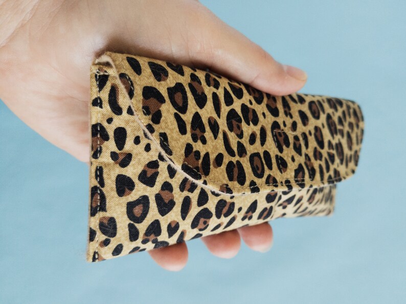 Puede incluir: Estuche para gafas con estampado de leopardo hecho de tela. El estuche se sostiene en una mano y tiene un cierre de solapa. El estuche presenta un fondo beige con manchas marrones y negras. Dise&ntilde;ado para guardar gafas.