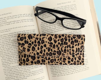 May include: Black-framed eyeglasses rest on an open book next to a leopard print glasses case. The case is rectangular with a curved flap closure. The book's pages are yellowed, and the background is a light blue.