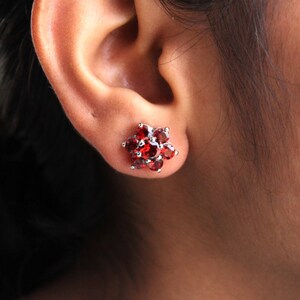 Peut inclure: Une paire de boucles d'oreilles clous en forme de fleur avec des pierres précieuses rouges. Les boucles d'oreilles sont en argent et présentent un groupe de pierres précieuses rouges en forme de fleur.